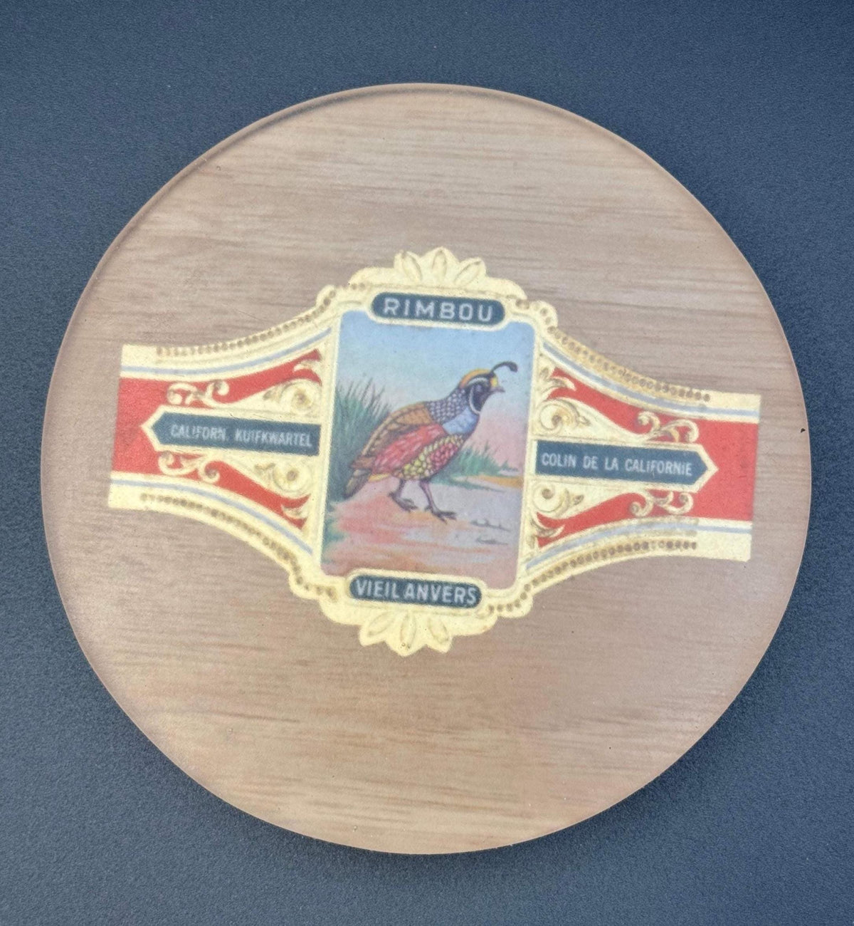 May include: A circular wooden coaster with a decorative label. The label displays a colourful illustration of a quail and the text "RIMBOU", "CALIFORN. KUIFKWARTEL", "COLIN DE LA CALIFORNIE", and "VIEIL ANVERS". vintage cigar, cigar coasters, cigar box art, antique cigar, whiskey coaster, bar drink coaster, cigar lounge decor, man cave decor, epoxy coasters, wood coasters, cigar gift, retro bar decor, classic bar decor
