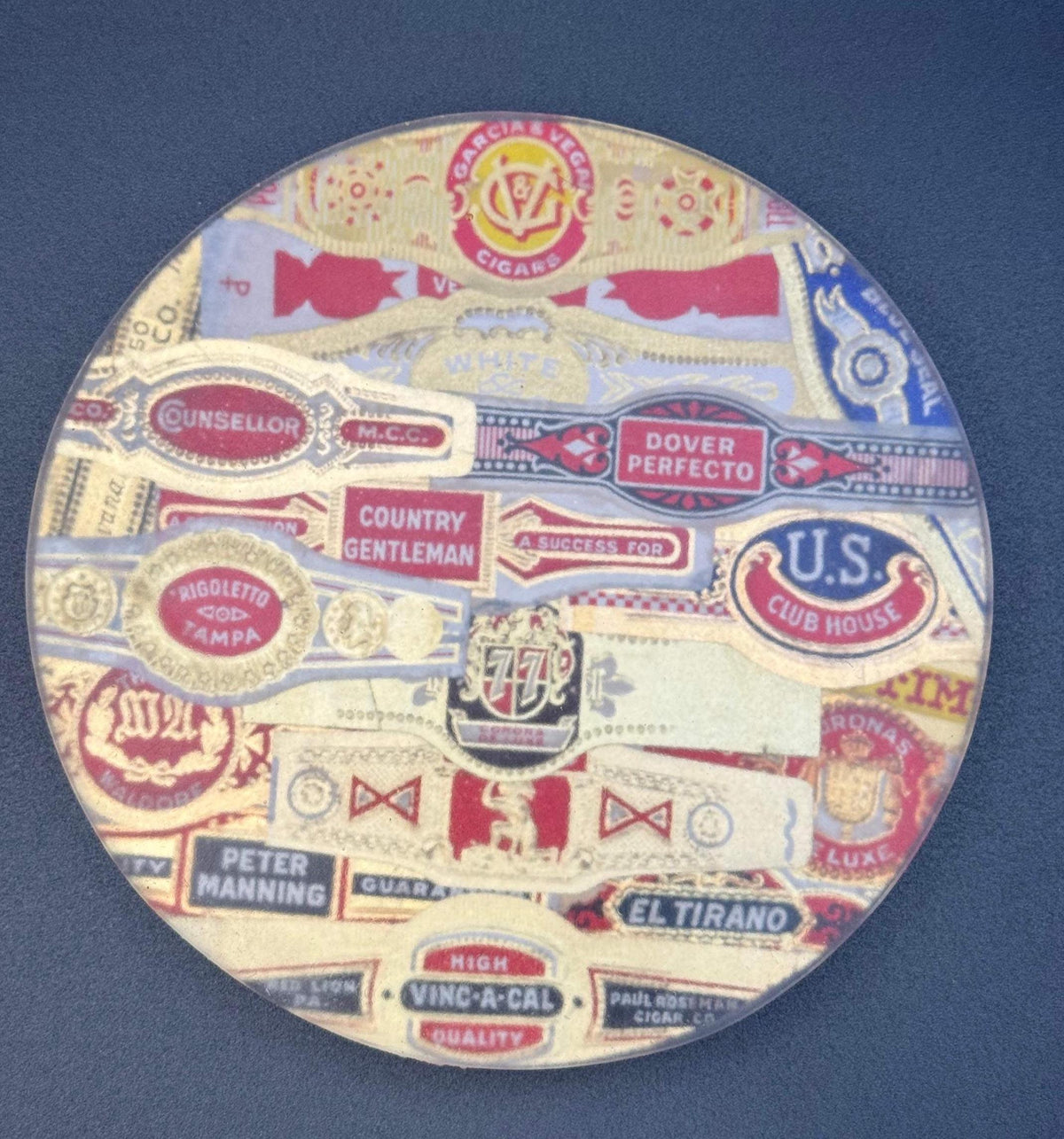 May include: Round coaster with a collection of vintage cigar labels. The labels are in various colours, including red, gold, blue, and white, with text such as "Garcia & Vega Cigars" and "U.S. Club House". A decorative item for a bar or home. vintage cigar, cigar coasters, cigar box art, antique cigar, whiskey coaster, bar drink coaster, cigar lounge decor, man cave decor, epoxy coasters, wood coasters, cigar gift, retro bar decor, classic bar decor